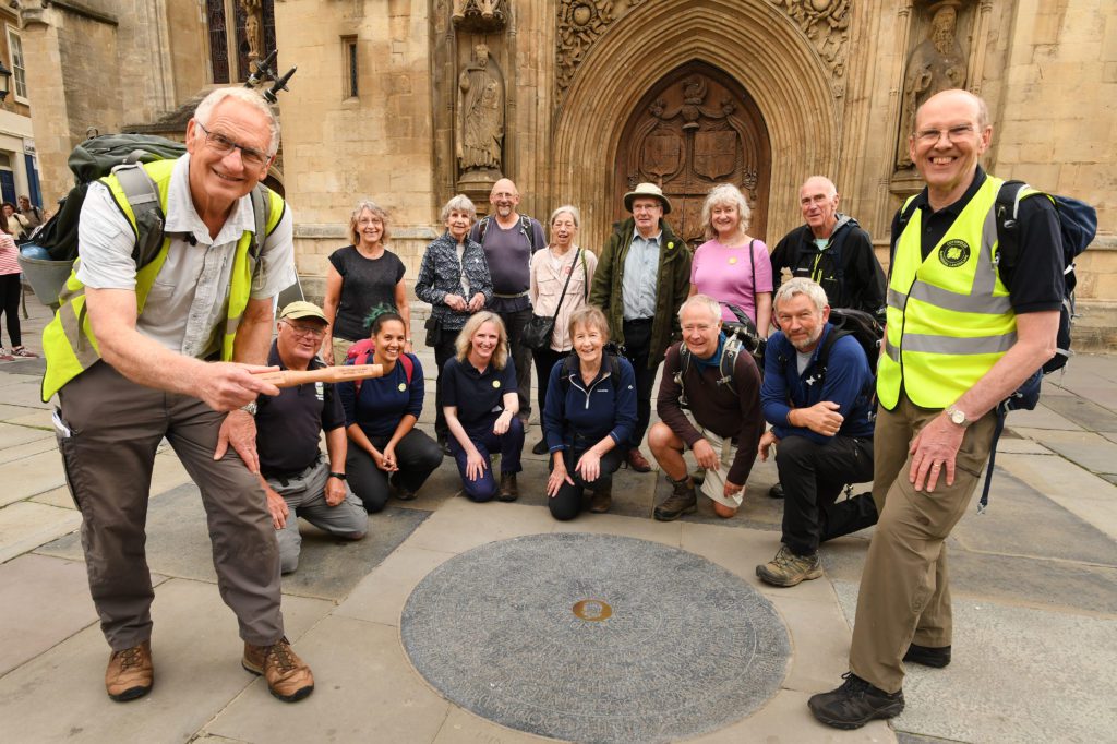 15 walkers infront of the west door of Bath Abbey, kneeling infront of the circular engraved Cotswold Way marker on the pavement area.