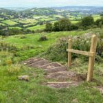 Completed mud steps reinforced with wood planks and handrail leading down the slope, and green rolling countryside fields beyond