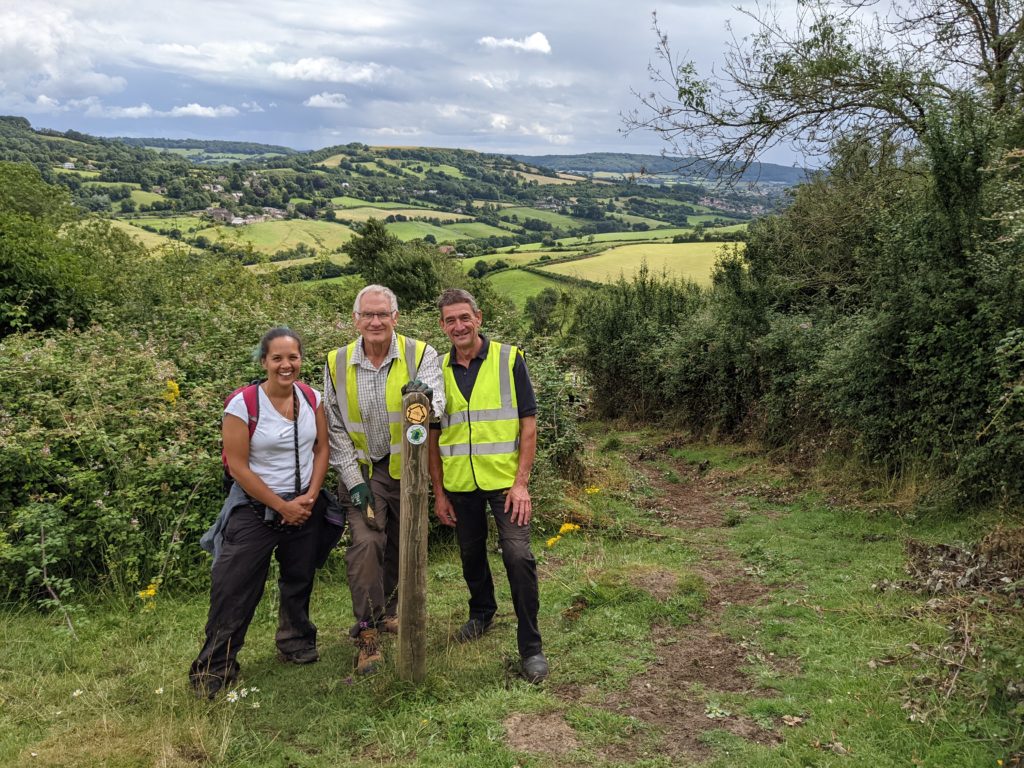 Nicole standing with two wardens at a marker post woth rolling green countryside hills and fields in the background