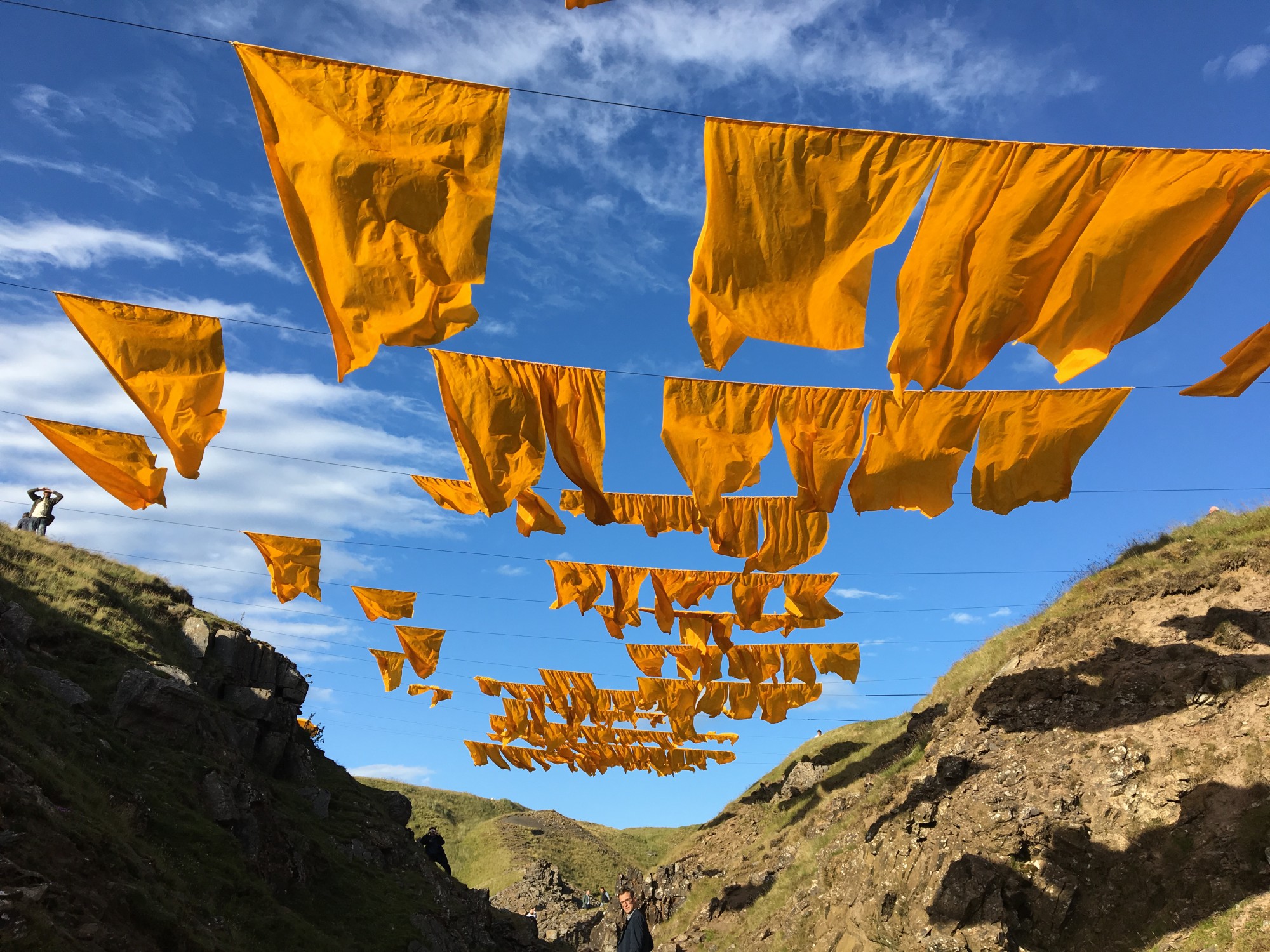 Large mustard yellow square flags suspended from horizontal lines flutter over grey rocky ravine with blue skies above