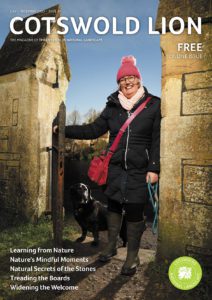 Image shows the front cover of Cotswold Lion magazine issue 49. A woman is shown wearing a winter parka coat, a cross body bag, and a pink bobble hat. She is pushing open a metal gate between two limestone gate posts. She is followed by a medium sized black dog. 