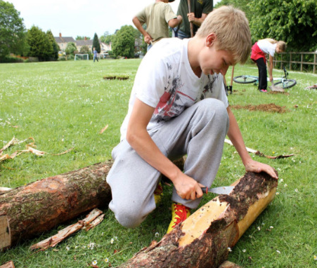 Image shows a young boy kneeling on grass chiselling a log.