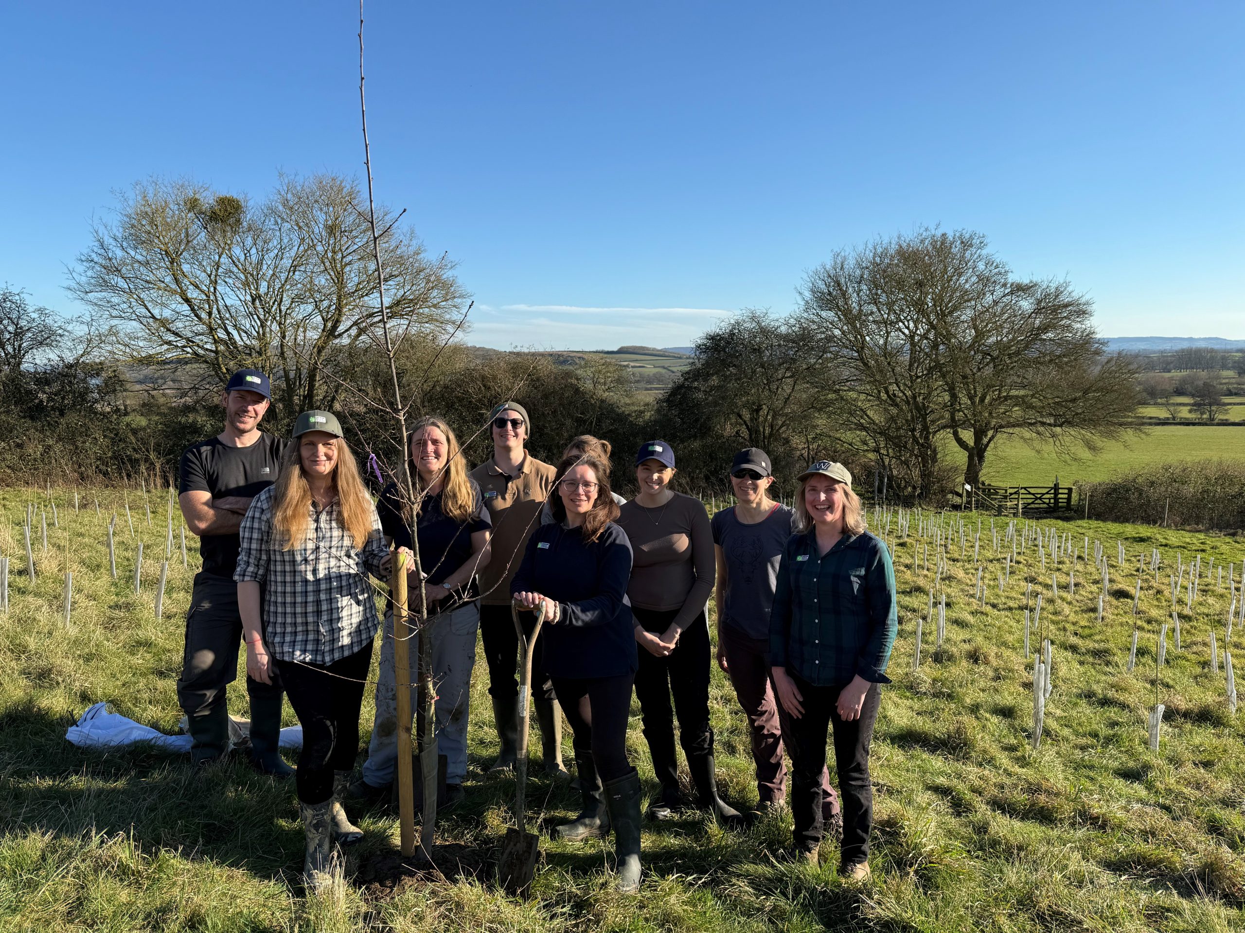 Image shows a group of CNL team members posing around a freshly planted tree, surrounded by other planted trees in a field. the sky is blue and the sun is shining.