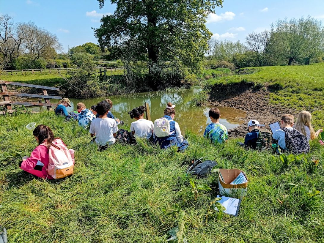 Image shows a group of children sitting on the grass by a river on a sunny day.