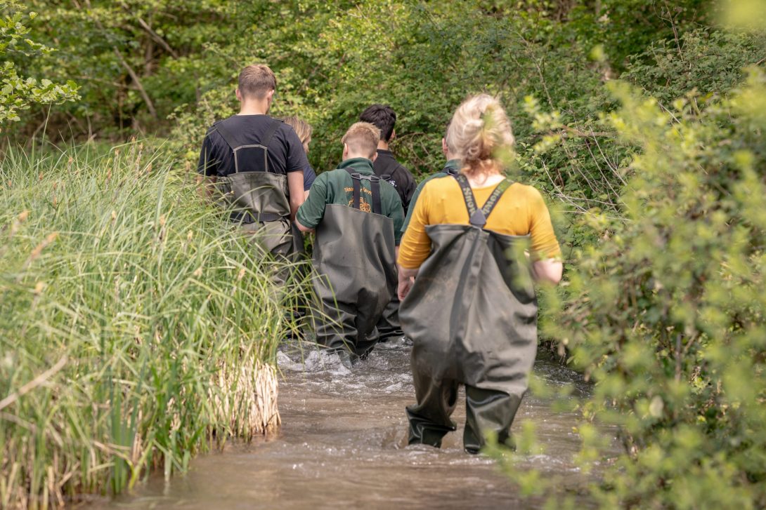 Image shows a small group of people dressed in waders walking through a river. Image by Tea Smart