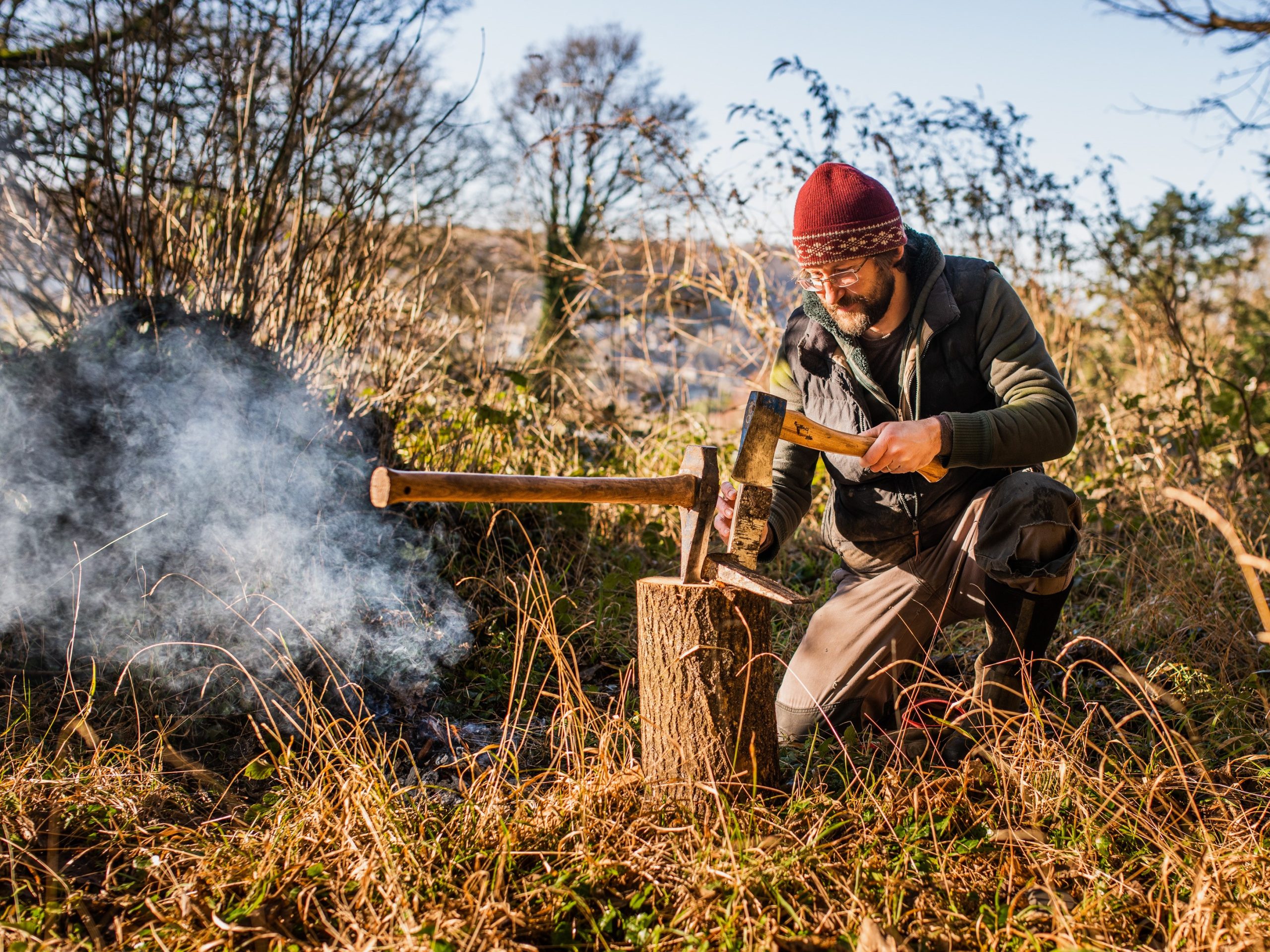 Photo shows a man crouching near a campfire, cutting kindling with a hatchet.
